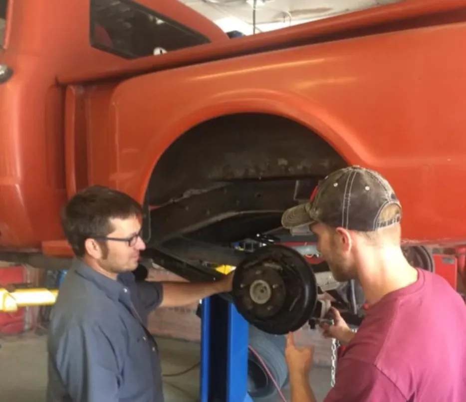 Two Star Auto Body technicians working on the wheel hub of an orange pickup on the lift